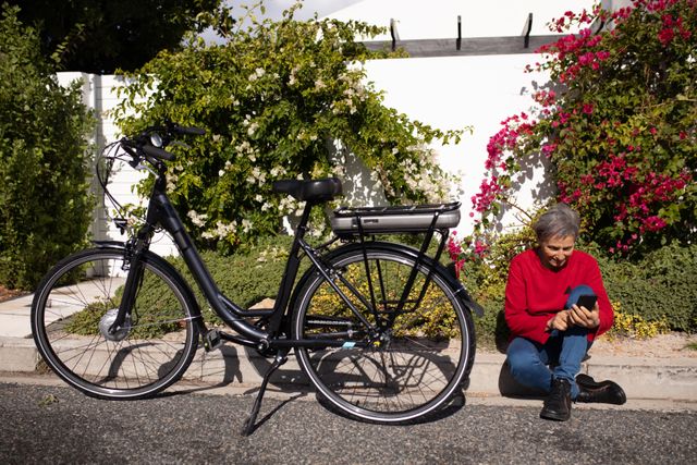 Senior woman sitting on curb using smartphone next to bicycle on a sunny day. Ideal for themes related to active aging, technology use among seniors, urban lifestyle, and outdoor leisure activities.