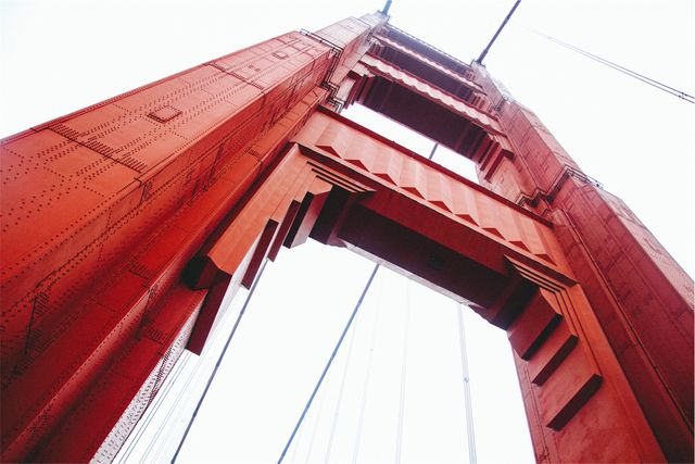 Perspective shot looking up at iconic red suspension bridge tower and cables, with sky background. Ideal for travel posters, architecture presentations, engineering studies, and promotional materials emphasizing landmarks or urban infrastructure.
