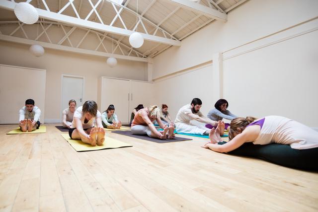 Group of people practicing pashimottanasana pose in a bright fitness studio with a trainer assisting. Ideal for promoting yoga classes, fitness programs, wellness retreats, and healthy lifestyle content.