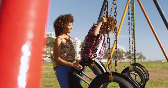 Mother and child enjoying playtime on a tire swing in a park on a sunny day. The image is perfect for illustrating family bonds, outdoor activities, parenting, and summer leisure. Suitable for use in blog posts, articles, or marketing materials relating to family time, parenting tips, lifestyle content, and outdoor activities.