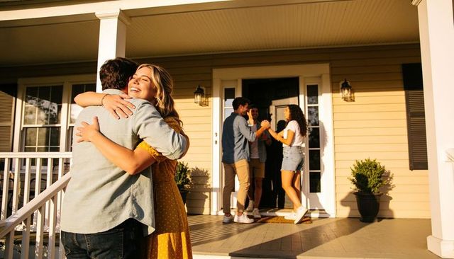 Young couple hugging on sunny front porch while friends welcome them at sunset