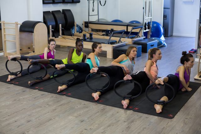 Group of women using Pilates rings during a workout session in a modern gym. The women are dressed in colorful sportswear and are positioned on yoga mats. This image can be used to promote fitness classes, women’s health, teamwork in sports, or advertisements for fitness equipment.