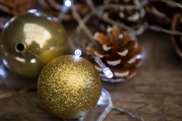 Golden Christmas baubles and pinecones on a wooden surface with festive lights create a warm and rustic holiday atmosphere. Ideal for use in holiday greeting cards, festive advertisements, seasonal blog posts, and social media content celebrating Christmas and winter holidays.