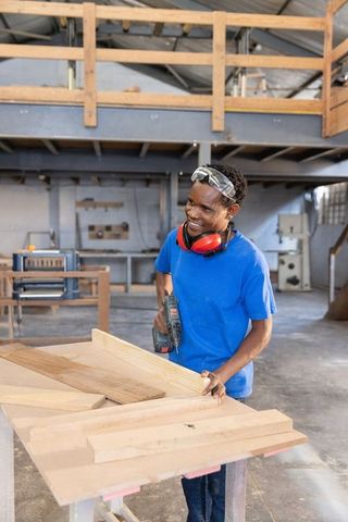 African american carpenter drilling wooden plank at workbench smiling with safety gear