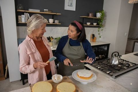 Mature women frosting layered cake together on marble kitchen island, home baking teamwork