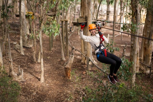 Woman enjoying an adventurous zip line ride through a forest, wearing a safety helmet and harness. Ideal for promoting outdoor activities, adventure sports, travel destinations, and active lifestyle content.