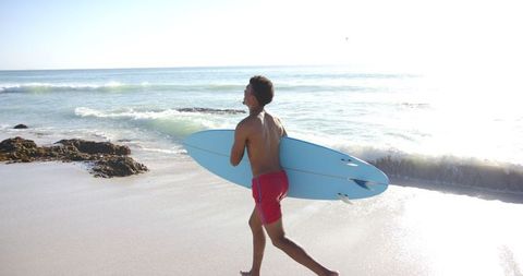 Young surfer walking on sunny beach with blue surfboard