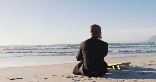 Senior african american man sitting with a surfboard at the beach. healthy outdoor leisure time by the sea.
