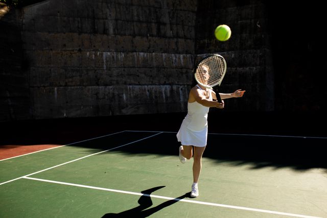 Front view of a Caucasian woman wearing tennis whites playing tennis on a sunny day, hitting a ball with a racket. 