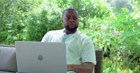 Man working on laptop in lush garden patio, preparing business report on wicker sofa