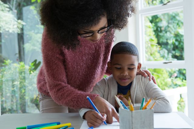 This image shows a mother helping her young son with his homework at home. It can be used to illustrate themes of parental support, education, family bonding, homeschooling, and childhood learning. The bright indoor setting with natural light highlights a nurturing and positive study environment.