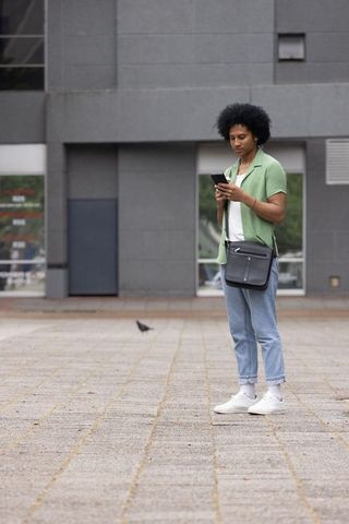 African american man checking phone on urban plaza wearing green shirt and crossbody bag