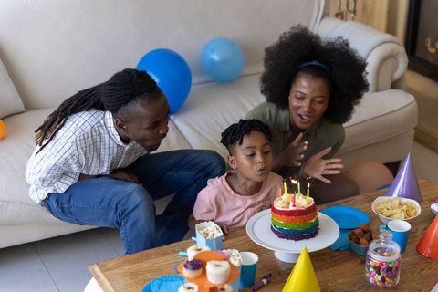 African american family celebrating child's birthday at home with rainbow cake and balloons