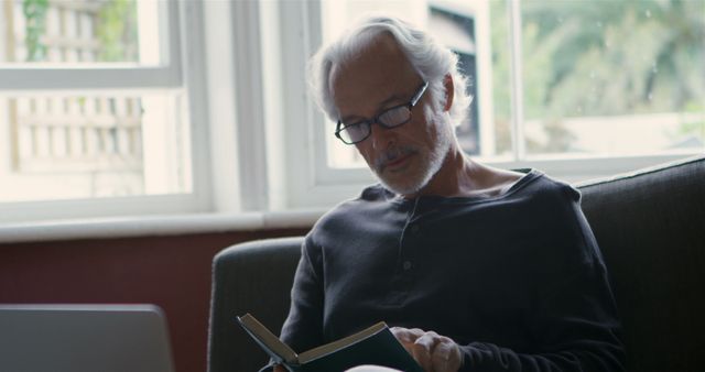 An elderly man with grey hair and glasses enjoying a quiet moment, reading a book while sitting on a couch next to a window. Perfect for illustrating themes of relaxation, tranquility, senior lifestyle, leisure activities, or indoor home environments.