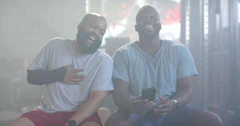 African american friends laughing on gym bench sharing smartphone after workout, casual mood