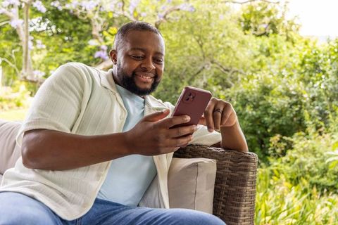 African man smiling and relaxing on wicker patio sofa using smartphone in lush garden