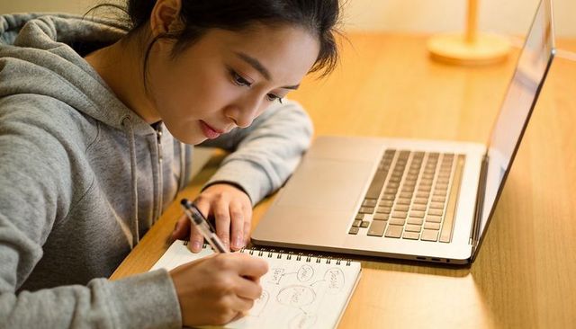 Focused student studying and writing notes at laptop on warm wooden desk