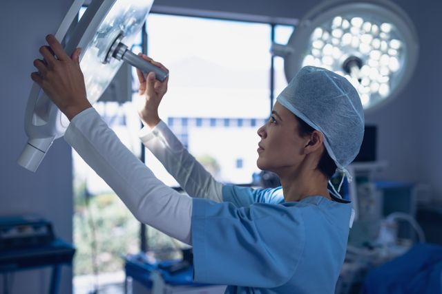 Female surgeon in blue scrubs and cap adjusting a surgical light in an operating room. Ideal for use in healthcare, medical, and hospital-related content. Can be used to depict surgical preparation, medical professionalism, and hospital environments.