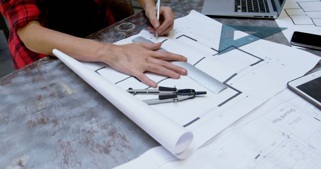 The image shows a person working on an architectural blueprint at a desk, using drafting tools such as a compass and ruler. This scene can be useful for websites or materials related to architecture, engineering, design, and technical drawing. It could also illustrate concepts of creativity, precision, and professional work environments.