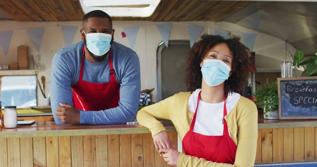 Two food truck staff members wearing face masks are standing behind a wooden counter, showing safety measures during pandemic. This can be used in articles or promotions related to food services, outdoor dining, health regulations, and small business operations.
