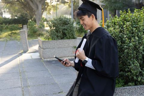 Asian graduate sitting on campus planter checking phone while holding diploma in cap and gown