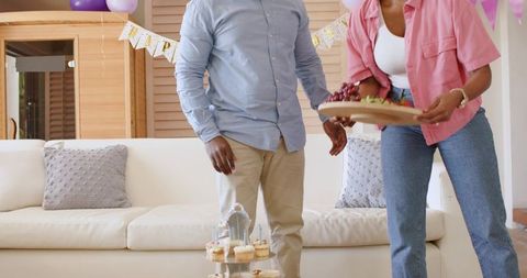 African american couple arranging festive living room party spread with cupcakes and grapes