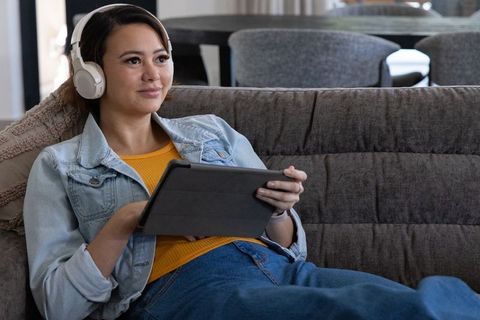 African american woman reclining on sofa listening with headphones and using tablet