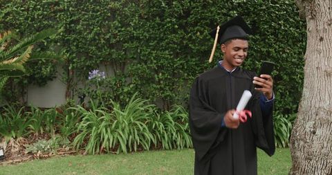 African American graduate celebrating on lawn while checking phone and holding diploma