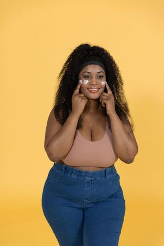 Smiling african american woman applying face cream wearing mauve tank top and denim
