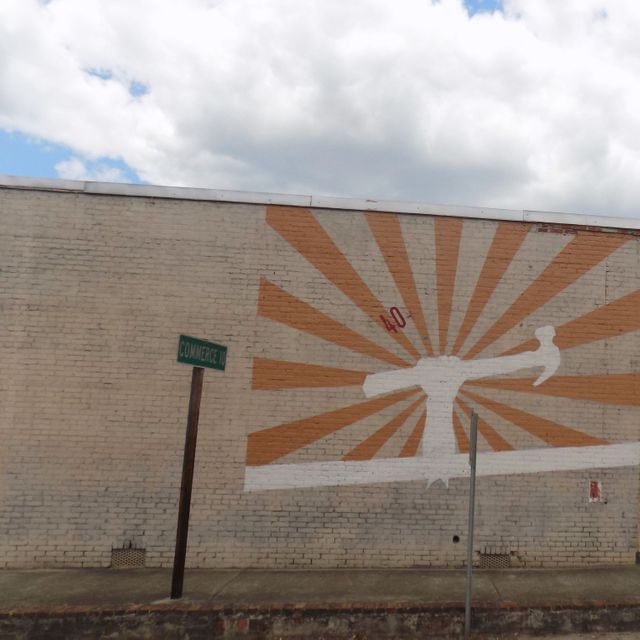 Brick wall adorned with a sunburst hammer mural under a partly cloudy blue sky, offering an urban, artistic atmosphere. Suitable for marketing materials related to street art, urban development projects, or creative city branding. Can also be used to represent innovation, craftsmanship, or commerce themes in promotional content.