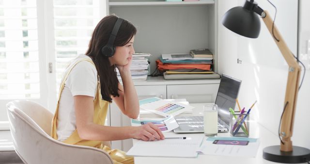 Young woman sitting at desk in home office, working on laptop while wearing headphones. Various office supplies and documents on desk with table lamp and shelves in background. Perfect for concepts of remote work, studying, freelancing, home office setup, and modern work environment.