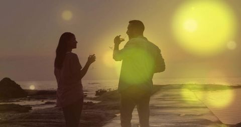 Couple enjoying sunset wine walk on rocky shore with golden lens flare