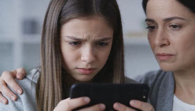 Mother comforting worried teenage daughter reading upsetting message on smartphone