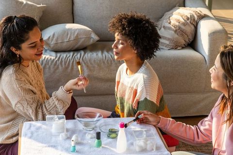 Diverse teenage friends enjoying diy spa day applying face masks and manicures at home