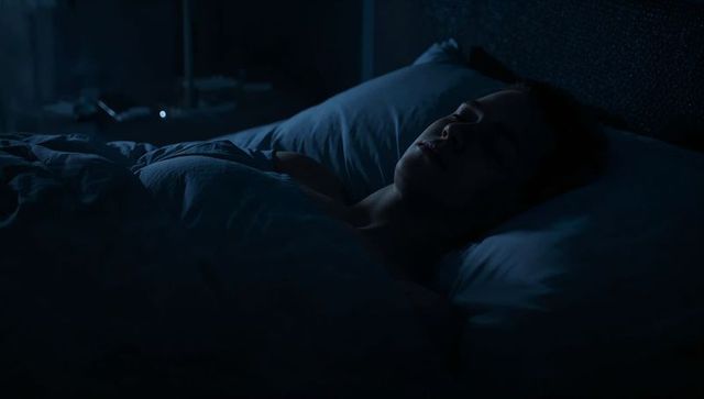 Shirtless man sleeping on back under rumpled duvet in moody blue bedroom