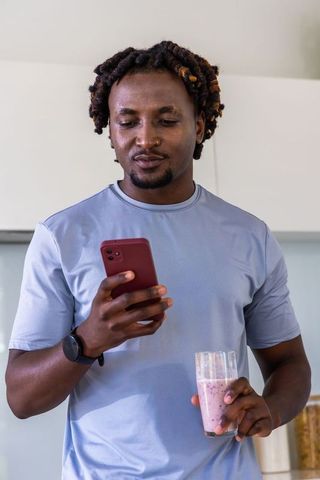Man checking phone while holding pink smoothie in kitchen wearing light-blue tee