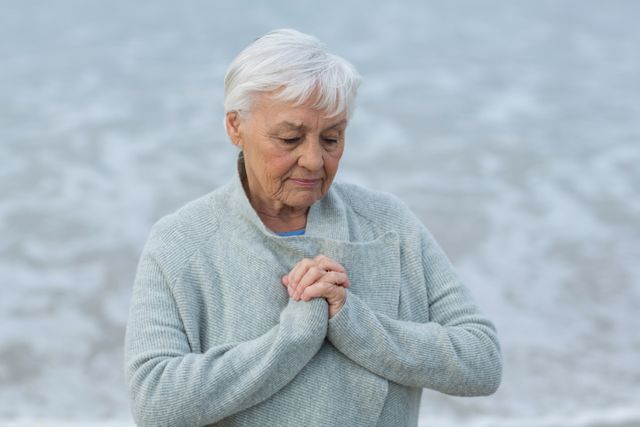 Senior woman with grey hair meditating on a beach, wearing a cozy sweater. Ideal for use in wellness, mental health, and aging gracefully themes. Perfect for promoting relaxation, mindfulness, and tranquility in natural settings.