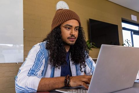 Young man typing on laptop in modern workspace wearing beanie and striped shirt