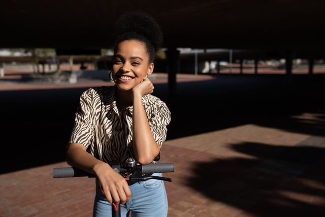 Portrait of a happy biracial woman enjoying free time in a city on a sunny day, standing in an urban park, smiling and leaning on an electric scooter, wearing blue jeans and zebra print shirt.