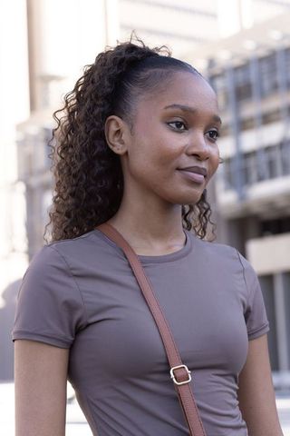 Confident african american woman standing in city plaza wearing mauve tee and leather bag
