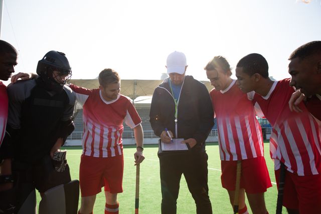 Multi-ethnic group of teenage male field hockey players, preparing before a game, standing in huddle with their coach holding a clipboard instructing. Sport game competition.