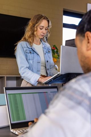 Woman standing at desk using laptop riser collaborating with coworker over spreadsheet