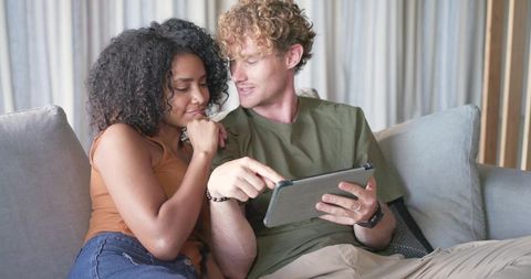 Interracial couple lounging on sofa browsing tablet together sharing digital moment