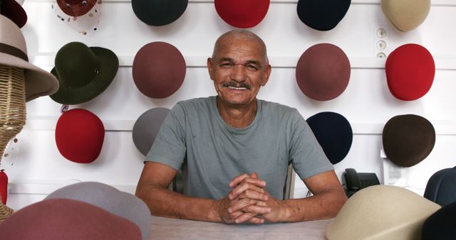 Happy senior bald biracial man with moustache sitting in showroom at hat shop, copy space. Millinery, hats, local business, retail, shop, work, production, tradition and craft, unaltered.