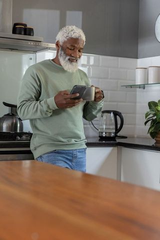 Senior african american man leaning on kitchen counter holding smartphone and coffee mug