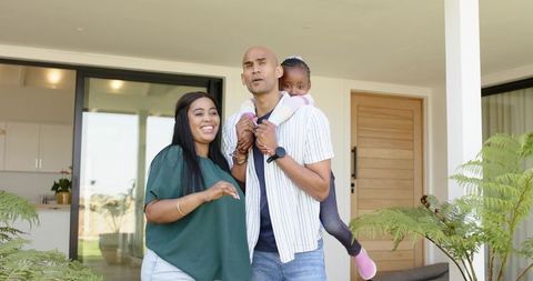 Diverse family enjoying sunny front porch, father piggybacking daughter while mother smiling