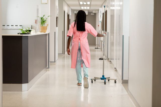 This image depicts an African American female patient walking down a hospital corridor while holding an IV stand. It can be used to illustrate themes of patient care, recovery, healthcare services, and hospital environments. Suitable for medical websites, healthcare brochures, and patient care articles.