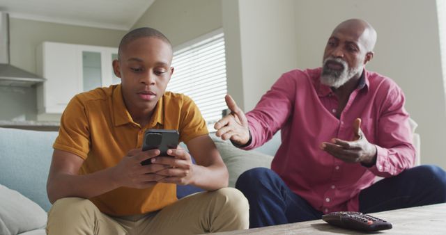 Father talking to teenage son who is using a smartphone while sitting in a living room. Picture highlights themes of communication, family relationship, and technology usage. Ideal for articles on parenting, father-son relationships, the impact of technology on family, and communication in the digital age.