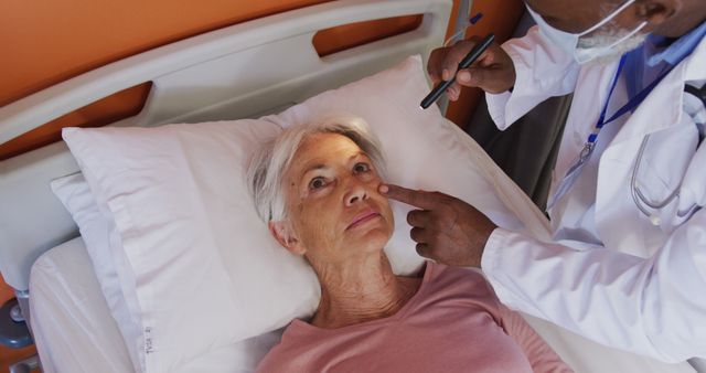 African american male doctor using penlight examining eye of senior caucasian female patient. Medicine, healthcare, lifestyle and hospital concept.