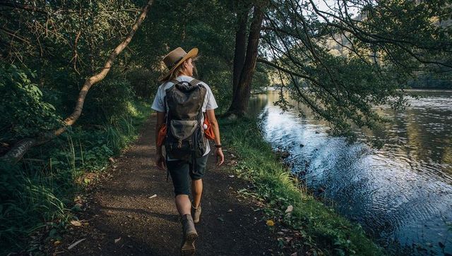 Solo hiker walking riverside trail under forest canopy with backpack and straw hat